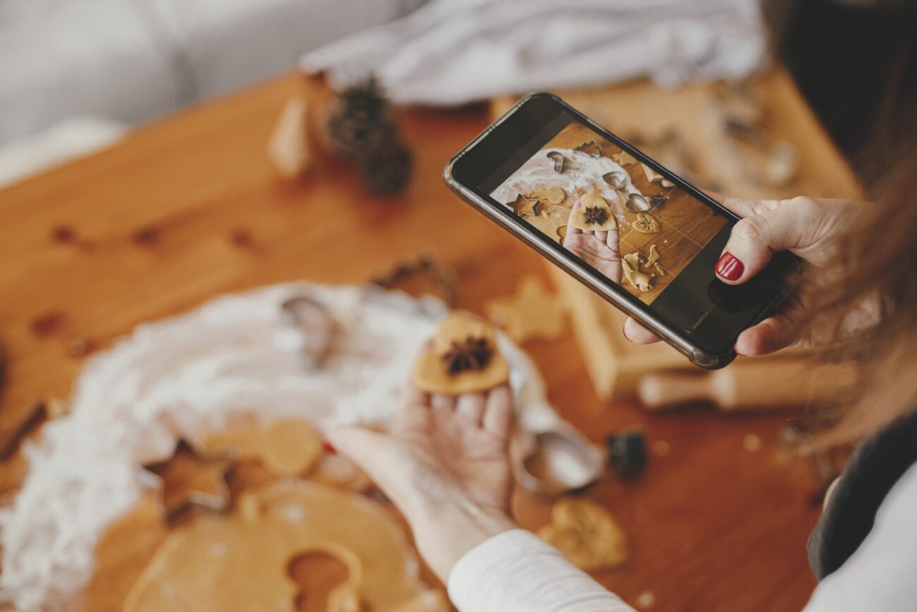 Woman holding a phone and taking photos of gingerbread Christmas cookies on a messy table flat lay. Instagram photo, social media, and blog. Holiday preparations